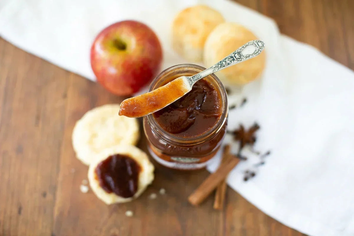 An open glass jar holds dark apple butter, with a spoon lifting a thick glossy portion above it. Apples, sliced bread, cinnamon sticks, and scattered spices are arranged on a white cloth over a wooden table.
