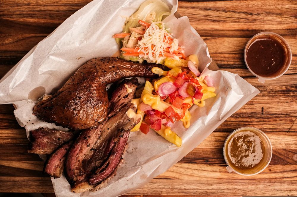 Close-up of assorted smoked barbecue meats on parchment paper over a wooden table with coleslaw and vegetables.