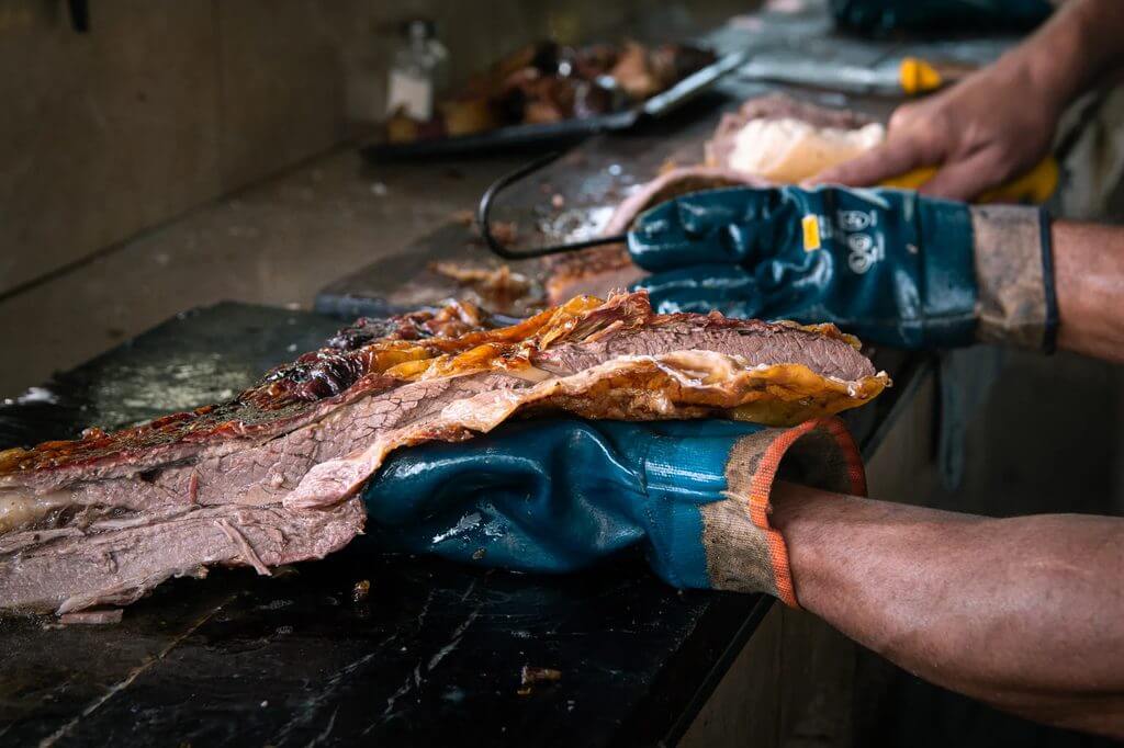 Butcher wearing gloves trims juicy brisket on a cutting board with a smoker in view