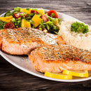 Grilled salmon fillets expertly seasoned with The Spice Lab Savory Whole Leaf 1 lb by The Spice Lab and a hint of BBQ seasoning, served alongside white rice, lemon wedges, and a fresh salad featuring mixed greens, tomatoes, and yellow bell peppers on a white plate atop a wooden table.