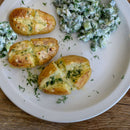 Three baked potatoes with melted cheese and herbs, including a sprinkle of The Spice Lab Dill Weed Whole, are on a white plate next to a cucumber salad with fresh dill and creamy dressing, set on a wooden table.