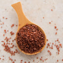 A wooden spoon filled with The Spice Lab Hawaiian Red Alaea Sea Salt Medium Grain crystals rests on a light, textured surface, with some of the brand’s coarse, reddish-brown salt scattered around, hinting at its earthy flavor.