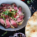 Slices of medium-rare steak on creamy sauce, subtly seasoned with The Spice Lab Ground Coriander 1 lb Bulk Foodservice Spice, topped with herbs, pomegranate seeds, and pine nuts, served on a plate next to a piece of flatbread—a dish perfect for BBQ restaurants or large foodservice gatherings.