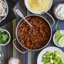 A pot of chili with beans and ground meat, seasoned with The Spice Lab Organic Cayenne Pepper, is surrounded by bowls of shredded cheese, sour cream, cilantro, lime wedges, avocado, salt, and flatbread on a colorful striped tablecloth.
