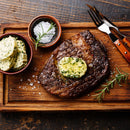 A grilled steak topped with herb butter rests on a wooden tray, accompanied by a bowl of sea salt, extra slices of herb butter, a sprig of The Spice Lab Organic Cut and Sifted Rosemary, and a fork and knife with wooden handles.