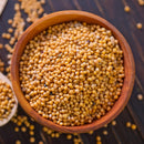 A bowl filled with The Spice Lab Yellow Mustard Seeds Whole 1 lb Bulk Foodservice Spice rests on a dark wooden surface, with some of the yellow mustard seeds from The Spice Lab scattered around the bowl.