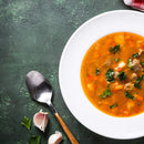 A bowl of vegetable and meat soup garnished with parsley on a white plate, infused with The Spice Lab Organic Smoked Paprika for a smoky-sweet flavor, is served alongside a spoon, garlic cloves, and parsley on a green textured surface.