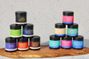Twelve spice jars with colorful labels are stacked on a wooden board against a speckled background. Perfect for enhancing classic Tuscany cuisine, World Spice Tuscan Rosemary Salt and other spices from World Spice—Long Pepper, Chorizo Bomb, Fenugreek Leaf, Smoked Salt, Tikka Masala, and more—pair beautifully with regional wines and a drizzle of extra virgin olive oil.