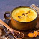 A rustic brown cup filled with The Spice Lab Golden Milk Turmeric Tea Superfood Premium Powder, featuring a rich golden hue, is topped with aromatic spices and rests on a matching saucer. A wooden spoon, star anise, and turmeric slices are arranged beside it on a burlap cloth.