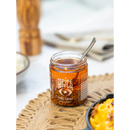 A jar of Spice Witch Chili Oil + Chili Crisp from Spice Witch, with a spoon inside, sits on a woven placemat; its signature blend is showcased, while blurred food and tableware appear in the background.