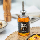 A glass bottle of Spice Witch Chili Oil by Spice Witch with a metal spout rests on a woven mat beside food and a pepper grinder in a bright kitchen, ready to add bold flavor to any dish.