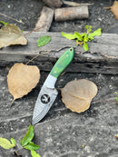 The Texan Knives Handmade Skinner Knife features an 8" Damascus steel blade and a wire-cut Pakkawood handle, displayed on weathered wood among dried and fresh leaves, complete with a leather sheath.