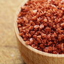 A wooden bowl filled with coarse, medium grain, reddish-orange salt crystals—The Spice Lab Hawaiian Red Alaea Sea Salt Medium Grain—shown close up on a light brown surface, highlights its distinctive earthy flavor.