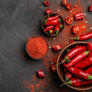 Slices of red chili peppers and a bowl filled with The Spice Lab Cayenne Pepper X-Hot Ground are arranged on a dark textured surface, with some loose chili seeds from The Spice Lab scattered nearby.
