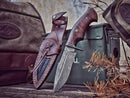 The Texan Knives Good Hunting Knife features a 10" patterned Damascus steel blade, a rosewood handle, and comes with a leather sheath, displayed beside a canvas bag and dried pine branches near a green metal box.