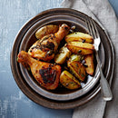 A plate featuring roasted chicken drumsticks seasoned with The Spice Lab Ground Coriander 1 lb Bulk Foodservice Spice and golden potato wedges, garnished with herbs and seeds, sits on a folded napkin with a fork and knife beside it—making it an ideal choice for BBQ restaurants aiming to serve up delicious foodservice-size portions.