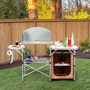 DirectDeals Foldable Outdoor BBQ Portable Grilling Table With Windscreen Bag set up on a lawn, with storage shelves, paper towel holder, drinks, and utensils; wooden fence and trees in background.
