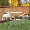 The DirectDeals Foldable Outdoor BBQ Table Grilling Stand is set up on grass with food, drinks, and utensils—ideal as a camping cook station, shown with a wooden fence and trees in the background.