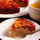 A slice of banana upside-down cake, subtly enhanced with aromatic spice and The Spice Lab Ground Star Anise for a delicate, licorice-like flavor, sits on a white plate with a fork, next to a cup of tea and the remaining cake in the background.