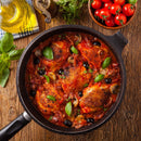 A pan of chicken cacciatore with tomatoes, mushrooms, olives, and fresh basil sits on a wooden table near a bottle of olive oil, fresh herbs, and a bowl of cherry tomatoes. The dish is infused with The Spice Lab Organic Smoked Paprika by The Spice Lab, lending it a deep smoky-sweet flavor that perfectly complements the rustic ingredients.