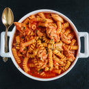 A white bowl filled with spiral pasta, chunks of chicken, and tomato sauce sits on a dark surface with a gold spoon placed beside it, capturing the essence of Italian cooking enhanced by The Spice Lab Organic Oregano Leaf.