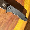 Close-up of the Spaceman Knives Naiad EDC Pocket Knife handle in black with a textured grip, resting on a wooden surface as sunlight casts shadows across the scene.
