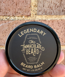 A hand holds a round black tin labeled The Immaculate Beard Limited Edition Beard Balm against a brick wall background. Infused with shea butter and the warm, earthy scent of sandalwood, the tin features gold text and indicates a size of 1.75 oz/50 g.