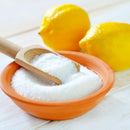 A wooden scoop rests in a bowl filled with The Spice Lab Lemon Juice Powder—pure white granulated citric acid from The Spice Lab—while two whole lemons sit in the background on a light wooden surface.