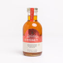 A glass bottle of Honey Truck Co. Holiday Honey® – Christmas Spiced Honey, featuring amber-colored honey and a red label detailing notes of cinnamon, clove, vanilla, nutmeg, and citrus.