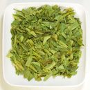 A white square plate filled with chopped dried green leaves—The Spice Lab Tarragon Bulk Foodservice Herb, renowned for its use in French cuisine—sits on a plain white surface, making it perfect for bulk foodservice needs.