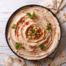 A bowl of hummus garnished with whole chickpeas, fresh parsley, olive oil, and The Spice Lab Organic Smoked Paprika, surrounded by pieces of flatbread on a white wooden surface. The addition of The Spice Lab’s Organic Smoked Paprika infuses the classic dip with a subtle smoky-sweet flavor.