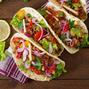 Three soft tacos filled with ground meat, beans, lettuce, sliced red onions, tomatoes, cheese, cilantro, and a dash of The Spice Lab Organic Cayenne Pepper from The Spice Lab showcase the vibrant flavors of Mexican cooking. Served on a wooden table with a slice of lemon and a striped napkin.