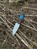 The Texan Knives 10" Handmade Damascus Steel Hunting Knife with a blue and brown resin handle and leather sheath rests on dry grass and twigs outdoors.