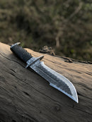 The Texan Knives Best Bowie Knife features a 13" handmade Damascus steel blade and resin horn handle, shown resting on a weathered log with a blurred natural background. Comes with a leather sheath.