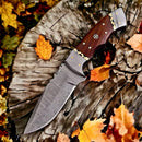 The Texan Knives Small Bushcraft Knife, featuring a 9" Damascus steel blade, rosewood handle, and leather sheath, sits on a weathered tree stump amid vibrant autumn leaves.