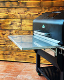 A Pimp My Grill® Stainless Steel Shelf Extension / Sleeve for Green Mountain Grills Jim Bowie / Peak attached to the front of a black pellet grill.