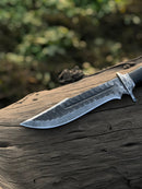 The Texan Knives Best Bowie Knife features a 13" handmade Damascus steel blade with an intricate pattern, resin horn handle, and leather sheath, showcased on a weathered wooden log with blurred greenery in the background.