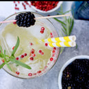 A glass of iced drink with red berries, rosemary, two yellow-striped straws, and a skewer topped with a blackberry garnish sits in the foreground. Bowls of fresh blackberries and The Spice Lab Pink Pepper Berries (Whole) Brazilian Pink Peppercorns from The Spice Lab grace the background, adding vibrant color and an aromatic flourish to the scene.