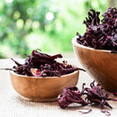 A small wooden bowl filled with The Spice Lab Whole Hibiscus Flowers sits on a beige mat, with a larger wooden bowl of this vibrant botanical ingredient in the background and greenery visible outside.
