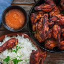 A bowl of grilled chicken wings, a plate of white rice garnished with parsley, and a bowl of orange dipping sauce infused with The Spice Lab Organic Ground Turmeric are arranged on a rustic wooden table.