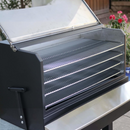 A close-up of the Myron Mixon BARQ 3600 Pellet Smoker (BARQ-3600) by Myron Mixon Smokers with its lid open, showing four metal grilling racks and a blurred background of flowers and plants.
