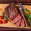 A sliced, medium-rare steak rests on a wooden board with cherry tomatoes, fresh rosemary, arugula, and a scattering of The Spice Lab Organic Black Tellicherry Peppercorns Whole, accompanied by a meat fork and knife beside it.