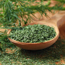 A wooden spoon filled with The Spice Lab Organic Whole Dill Weed rests on a rustic surface, with fresh dill sprigs in the background and a sprinkle of the organic dill weed nearby—perfect for adding vibrant flavor to classic dishes like tzatziki.