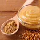A glass bowl of smooth mustard sits beside a wooden spoon filled with ground mustard powder, with The Spice Lab Yellow Mustard Seeds Whole 1 lb Bulk Foodservice Spice and yellow mustard seeds from The Spice Lab scattered across a wooden surface, creating a vibrant display of mustard seed spice.
