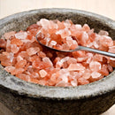 A metal spoon scoops The Spice Lab Himalayan Pink Salt Coarse Grain crystals from a round, rough stone bowl on a light wooden surface—perfect for use in your salt grinder.