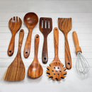 Eight wooden kitchen utensils from the Spice Wheel Acacia Wooden Spoon Set, along with one metal whisk, are arranged side by side on a white wooden surface. The set, crafted by Spice Wheel, includes spoons, spatulas, and a pasta server.