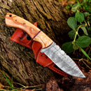 The Texan Knives Custom Handmade 10" Damascus Steel Hunting Knife features a patterned blade, olive wood handle, and comes with a leather sheath, displayed on a tree trunk surrounded by green leaves.