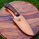 The Texan Knives Damascus Steel Hunting Knife – 10" with Rosewood Handle and Leather Sheath is displayed on a round, natural-grain wood board atop green grass.