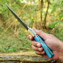 A hand holds the Texan Knives Custom Handmade Damascus Steel Hunting Knife, featuring a 10-inch blade and a blue resin handle, outdoors with blurred green foliage in the background; includes leather sheath.
