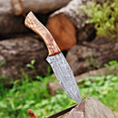 The Texan Knives Exquisite 10-Inch Damascus Steel Hunting Knife with a rosewood handle and leather sheath is embedded in a tree stump, with blurred logs and greenery in the background—an ideal gift for outdoorsmen.